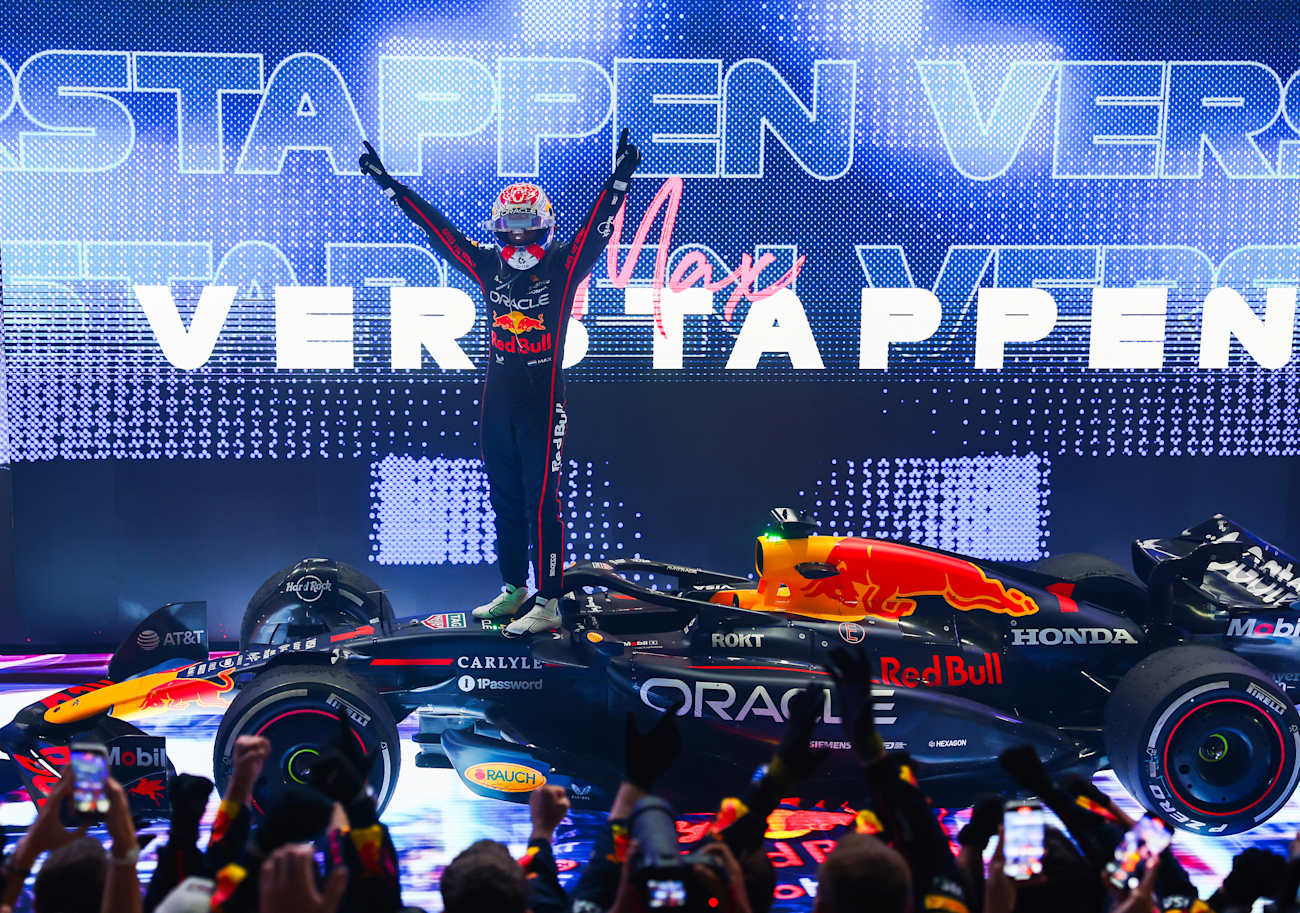 Formula 1 driver celebrating victory with a car on a stage, large 'Verstappen' text in the background - Ex Race Gear